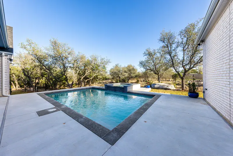 Pool with a dark stone edge beside the white brick house, Farmhouse Hill Country Retreat (RenovatePros project)