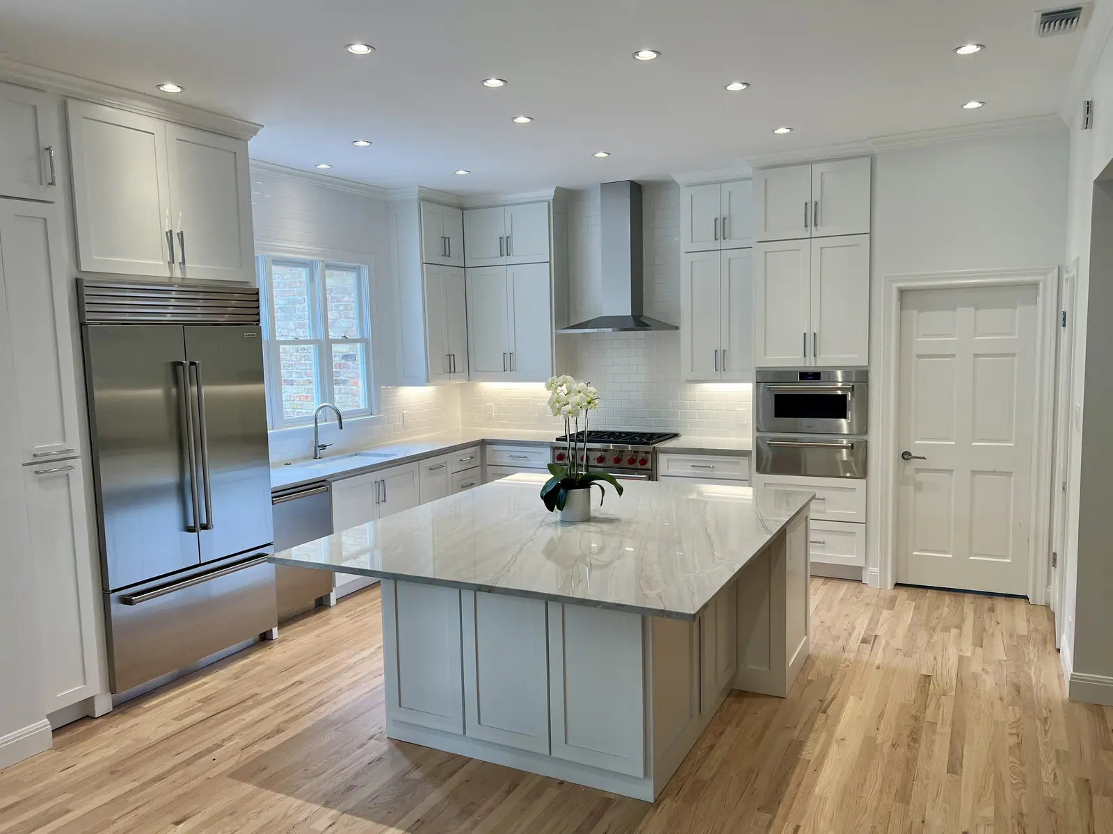 Remodeled kitchen with white shaker cabinets, a large island with a stone top, a stainless range hood and light wood floors, Rollingwood Remodel by RenovatePros